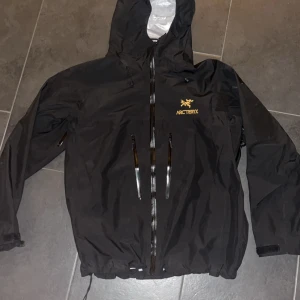 Svart vindjacka från Arc'teryx - Säljer en svart vindjacka från Arc'teryx med huva och dragkedja framtill. Jackan har två framfickor med dragkedjor och är perfekt för utomhusaktiviteter. Den är designad för att stå emot vind och regn. Storlek Xl men den e slim så ja som har m passar den bra 
