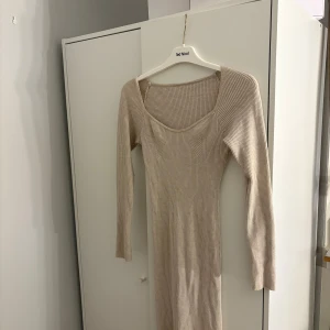 Beige ribbad klänning - Snygg beige ribbad klänning med lång ärm och en elegant v-ringad design. Perfekt för en stilren look. Klänningen har en slits som ger extra rörelsefrihet.