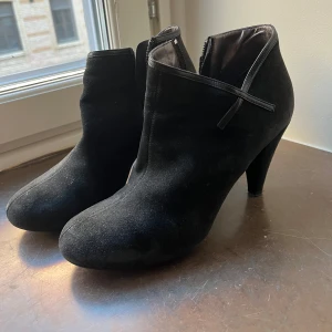 Svarta mocka boots med klack - Snygga svarta boots i mocka med klack och dragkedja på sidan. Perfekta för en stilren look. De har en spetsig tå och en diskret remdetalj.