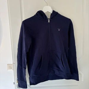 Mörkblå hoodie med dragkedja från Gant - Säljer en mörkblå hoodie från Gant med dragkedja. Hoodien har en klassisk design med Gant-logga på bröstet och ribbade muddar vid ärmslut och nederkant. Storlek: 158/164.