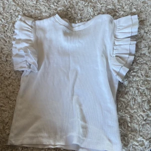 Vit ribbad topp från Gina Tricot - Säljer en söt vit ribbad topp från Gina Tricot med volangdetaljer på ärmarna. Perfekt för en stilren look. Toppen har en normal passform och är tillverkad i ett mjukt material.