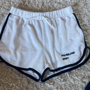 Vita shorts från Gina Tricot - Säljer ett par vita shorts från Gina Tricot med svart kant och snörning i midjan. Perfekta för sportiga aktiviteter eller en avslappnad dag. De har texten 'Philadelphia Sport' broderad på framsidan.