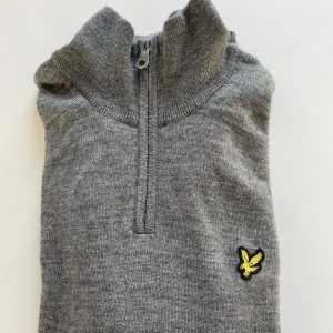Grå tröja med dragkedja från Lyle & Scott - Snygg grå tröja från Lyle & Scott med halv dragkedja och ribbad krage. Den har ett gult logomärke på bröstet. Perfekt för en stilren och avslappnad look.