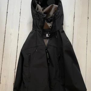 Svart CP Company Shell Jacket  - Säljer en stilren svart vindjacka från CP company i väldigt bra skick. Jackan har en normal passform och är perfekt för blåsiga och regniga dagar. Den har praktiska fickor med dragkedja och justerbar snörning i huvan.