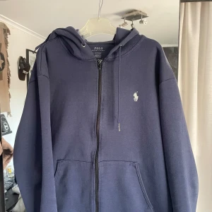 Mörkblå hoodie från Ralph Lauren - Säljer en snygg mörkblå hoodie från Ralph Lauren med dragkedja och huva. Den har en klassisk design med det ikoniska logotypen broderad på bröstet. Perfekt för en avslappnad stil.