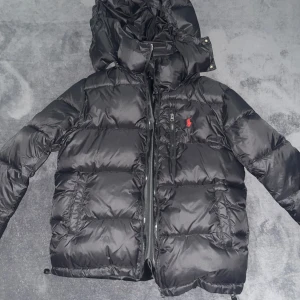 Svart dunjacka från Ralph Lauren - Säljer en snygg svart dunjacka från Ralph Lauren med en röd broderad logga på bröstet. Jackan har en praktisk dragkedja och en justerbar huva. Perfekt för kyliga dagar!