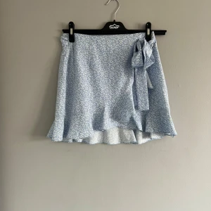 Blå blommig kjol från Shein - Säljer en söt blå blommig kjol från Shein. Kjolen har en omlott-design med en knytning i sidan och volangdetaljer i nederkant. Perfekt för en somrig look! 🌸Obs! Den har en lite svart fläck!