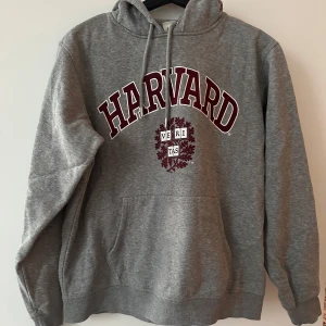 Grå hoodie med Harvard-tryck - Säljer en grå hoodie med Harvard-tryck i vinrött och vitt. Tröjan har en klassisk känguruficka och justerbar dragsko i huvan. Perfekt för en avslappnad stil eller som ett statement-plagg. 🐻