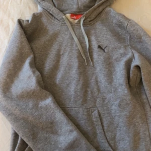 Grå hoodie från Puma - Säljer en grå hoodie från Puma med en klassisk känguruficka och justerbar huva. Perfekt för en avslappnad stil. Den har en liten Puma-logga på bröstet och är tillverkad i mjukt material för extra komfort.
