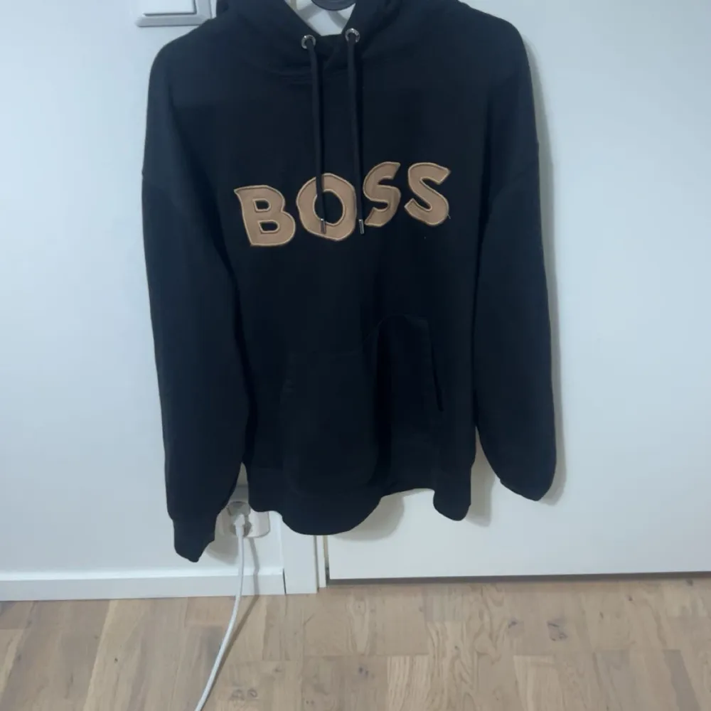 Säljer en svart hoodie från Boss med stor logga i beige framtill. Tröjan har en klassisk design med huva och dragsko samt en stor ficka på magen. Perfekt för en avslappnad stil.. Neuletakit & Villapaidat.