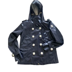 Marinblå regnjacka från Moncler med huva - Snygg marinblå regnjacka från Moncler med stora vita knappar och huva med dragsko. Jackan har dubbelknäppning framtill, fickor på sidorna och justerbara ärmslut. Perfekt för regniga dagar när du vill hålla stilen.