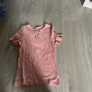 Rosa ribbad topp med snörning - Säljer en ljusrosa ribbad topp från Boohoo med korta ärmar och snygg snörningsdetalj vid halsen. Perfekt för dig som gillar en enkel men trendig stil. Toppen är mjuk och stretchig för extra komfort.