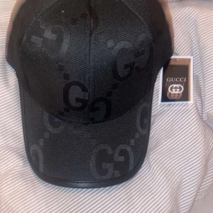 Svart keps från Gucci med GG-mönster - Snygg svart keps från Gucci med klassiskt GG-mönster över hela kepsen. Justerbar rem baktill och diskret logotyp på insidan. Perfekt accessoar för en trendig look.