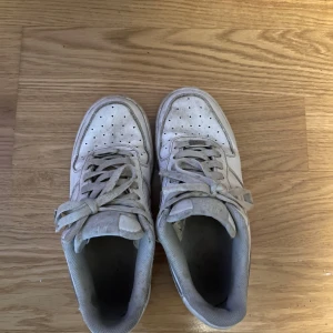 Vita Nike Air Force 1 sneakers - Klassiska vita Nike Air Force 1 sneakers med snörning och rund tå. Skorna har en platt sula och är tillverkade i syntetmaterial med detaljer i skinn. Perfekta för en streetwear-look.