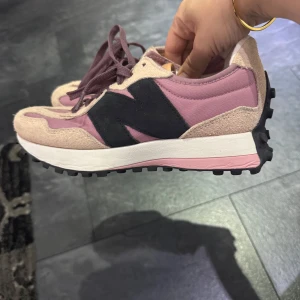 Lila och beige sneakers från New Balance - Säljer ett par snygga New Balance sneakers i lila och beige med svarta detaljer. Skorna har en kraftig sula med gummidobbar och klassisk snörning. Perfekta för dig som vill ha en cool och sportig stil.