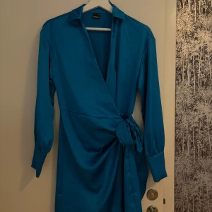 Blå omlottklänning med knytband - Säljer en snygg blå omlottklänning med knytband i midjan och långa ärmar med knappar vid ärmsluten. Klänningen har en elegant krage och är tillverkad i ett glansigt, mjukt material som ger en lyxig känsla.