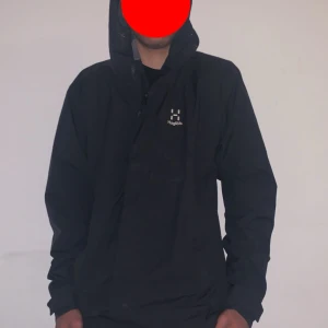 Svart vindjacka från Haglöfs - Säljer en svart vindjacka från Haglöfs med huva och dragkedja. Jackan har en stilren design med diskret logga på bröstet och är perfekt för blåsiga dagar. Tillverkad i syntetmaterial och passar dig som vill ha en lätt och funktionell jacka.