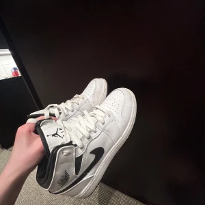 Nike Air Jordans i vitt och svart - Säljer ett par klassiska Nike Air Jordans i vitt med svarta detaljer och svart swoosh. Skorna har snörning, rund tå och platt sula. Perfekta för dig som gillar streetwear och vill ha en ikonisk sneaker i garderoben.