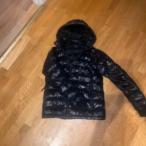 Svart dunjacka från Peak Performance - Säljer en svart dunjacka från Peak Performance med huva och dragkedja. Jackan har en glansig finish och är perfekt för kyligare dagar. Klassisk pufferstil med fickor framtill och diskret logga på insidan.