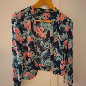 Blommig jacka från Gina Tricot - Säljer en tunn jacka från Gina Tricot med blommigt mönster i rosa, turkos och grönt på mörkblå botten. Jackan har dragkedja framtill och är långärmad. Perfekt för att liva upp din outfit!