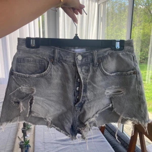 Grå slitna jeansshorts från Zara - Grå jeansshorts från Zara med råa, fransiga kanter och slitningar framtill. Klassisk femficksmodell med knappar i gylfen och hög midja. Perfekta för en avslappnad stil.