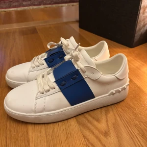 Valentino sneakers med blå detalj - Snygga vita sneakers från Valentino med en bred blå rem över snörningen och ikoniska nitar på hälen. Skorna har rund tå, platt sula och är tillverkade i skinn. Perfekta för dig som vill ha en stilren men unik look.
