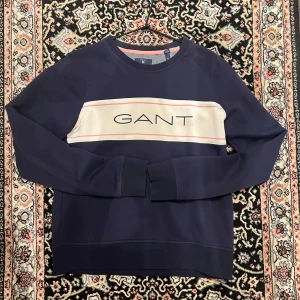 Marinblå sweatshirt från GANT - Säljer en marinblå sweatshirt från GANT med vit panel och rosa ränder över bröstet samt stor GANT-logga. Tröjan har rund halsringning och långa ärmar. Perfekt för en stilren och avslappnad look.