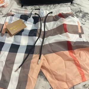 Rutig shorts från Burberry - Snygga rutiga shorts från Burberry i beige, vit, svart och röd. De har elastisk midja med svart snörning och klassiskt Burberry-mönster. Perfekta för sommaren och riktigt trendiga.