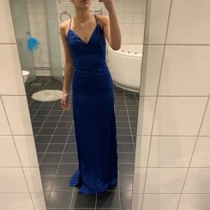 Blå balklänning med slits - Säljer en elegant blå lång balklänning med smala axelband som knyts vid halsen och djup v-ringning. Klänningen har en slits på ena sidan och figurnära passform, perfekt för balen. Materialet har en lätt glansig yta som ger en lyxig känsla. Passar xs och s 
