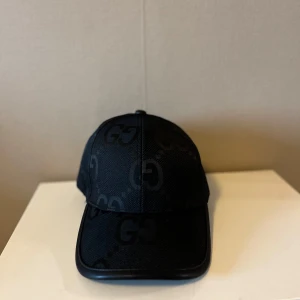 Svart keps från Gucci med GG-mönster - Säljer en svart keps från Gucci med diskret GG-mönster över hela kepsen. Justerbar rem baktill för perfekt passform. Snygg och stilren accessoar som lyfter vilken outfit som helst.