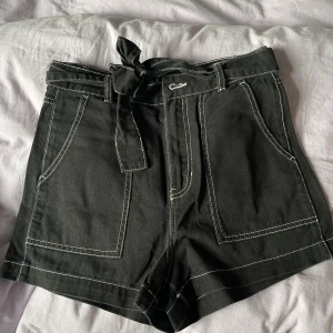 Svarta shorts med knytband och kontrastsömmar - Svarta shorts med vita kontrastsömmar och breda fickor fram och bak. Knytband i midjan och bälteshällor. Högmidjade och i ett lite kraftigare tyg, perfekt för en cool och avslappnad stil.