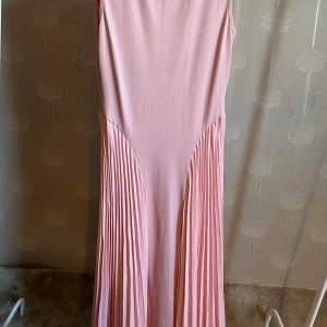 Ljusrosa långklänning - Säljer en elegant ljusrosa långklänning med plisserad kjol. Klänningen är ärmlös och har en figurnära överdel som övergår i en vackert veckad kjol. Aldrig använt, saknar prislapp