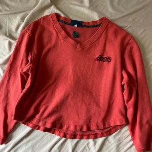 Röd croppad långärmad tröja från Asics - Säljer en röd långärmad tröja från Asics med broderad logga på bröstet. Tröjan har v-ringad hals och är croppad i modellen. 