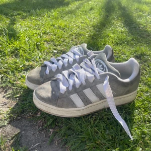 Adidas Campus grå sneakers - Säljer ett par grå Adidas Campus sneakers med vita ränder och vit sula. Skorna har klassisk snörning och är tillverkade i mocka med detaljer i syntet. Perfekta för dig som gillar en stilren och tidlös look.