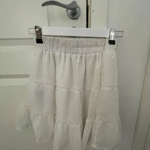 Vit volangkjol med prickigt mönster - Säljer en söt vit kjol med volanger och diskret prickigt mönster. Kjolen har resår i midjan och är lätt och luftig, perfekt för varmare dagar. Passar dig som gillar romantisk och feminin stil. Skriv för fler bilder och pris går att diskutera!! Vet ej vart den är ifrån