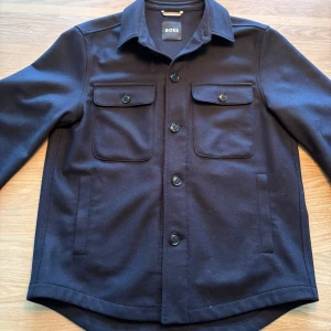 Marinblå overshirt/jacka  från BOSS - Stilren svart overshirt/jacka från BOSS. Jackan har avslappnad passform, klassisk krage, knäppning framtill. Perfekt för våren. Den är i storlek 48 och sitter relaxed fit på mig som är 177cm. Den är i nyskick, använd endast 1 gång.