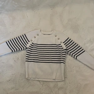 Vit och marinblå randig stickad tröja från Tommy Hilfiger - Säljer en vit stickad tröja från Tommy Hilfiger med marinblå ränder och dekorativa silverfärgade knappar på axlarna. Tröjan har lång ärm och ribbade detaljer vid hals, ärmslut och nederkant. Perfekt för en stilren och klassisk look.