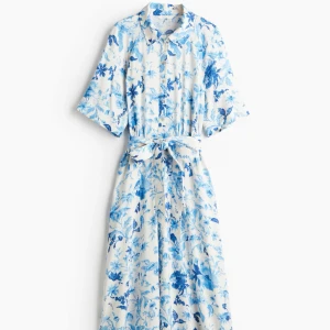 Blåvit blommig skjortklänning med knytband - Säljer en somrig blåvit skjortklänning med blommigt mönster och knytband i midjan. Klänningen har korta ärmar, krage och knappar framtill. 