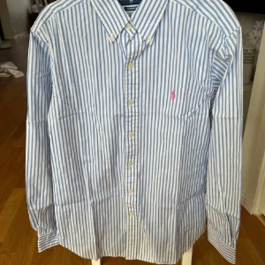 Blåvit randig skjorta från Ralph Lauren - Klassisk blå och vit randig skjorta från Ralph Lauren med button-down krage och broderad rosa logga på bröstet. Skjortan har lång ärm och knäppning framtill. Perfekt för en stilren look.