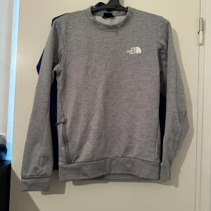 Grå sweatshirt från The North Face - Säljer en stilren grå sweatshirt från The North Face med rund halsringning och diskret logga på bröstet. Tröjan har långa ärmar och är tillverkad i mjukt material, perfekt för vardagsbruk eller träning.