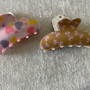 Färgglada hårklämmor med hjärtmönster - Två stora hårklämmor i plast, en i ljusrosa med hjärtan i rosa, lila och gult, och en i beige med små rosa hjärtan. Perfekta för att sätta upp håret med en söt touch. Ny 