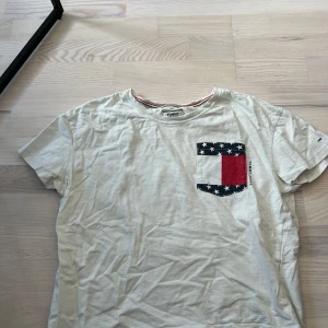 Vit t-shirt med stjärnmönstrad ficka från Tommy Jeans - Vit t-shirt från Tommy Jeans med en bröstficka i svart, vitt och rött med stjärnmönster och logga tryckt vid sidan. Klassisk rund halsringning och korta ärmar. Perfekt för en avslappnad och cool stil.