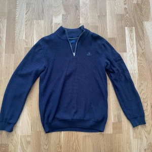 Mörkblå halvzip tröja från GANT - Säljer en mörkblå tröja från GANT med halv dragkedja och diskret strukturmönster. Tröjan har ribbade muddar och krage, samt GANT-logga broderad på bröstet. Perfekt för lager-på-lager och stilren look.