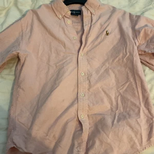 Ljusrosa skjorta från Ralph Lauren - Säljer en klassisk ljusrosa skjorta från Ralph Lauren med button-down krage och broderad logga på bröstet. Skjortan har långa ärmar och vita knappar framtill. Perfekt för en stilren look!