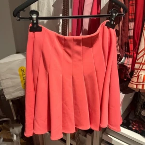 Rosa kortkjol med veck - Säljer en snygg korallrosa kortkjol med markerade veck och dragkedja i sidan. Kjolen har en lätt utsvängd form och är tillverkad i ett mjukt, slätt syntetmaterial. Perfekt för dig som vill ha en färgklick i garderoben!