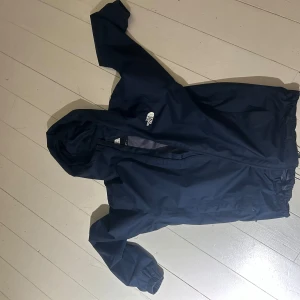 Mörkblå vindjacka från The North Face - Säljer en mörkblå vindjacka med huva från The North Face. Jackan har dragkedja framtill, elastiska muddar och logga på bröstet och ryggen. Perfekt för blåsiga dagar och enkel att packa ner i väskan. Knappt använt och inga defekter 