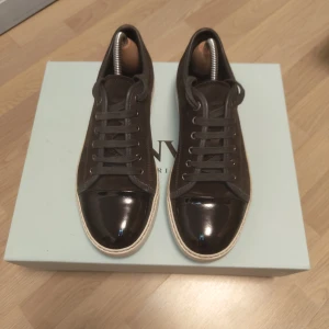 Bruna sneakers med lackad tå - Säljer ett par bruna sneakers från Lanvin med snygg lackad tå i svart och grå snörning. Skorna har vit sula och är tillverkade i en kombination av mocka och läder. Perfekta för dig som vill ha en stilren och exklusiv look. Skorna är storlek 41 men funkar även till 43
