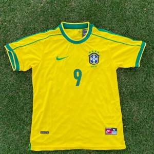 Brasilien World Cup Hemma fotbollströja Ronaldo 9 - Klassisk gul Brasilien fotbollströja med gröna detaljer, nummer 9 och Ronaldo på ryggen. Nike-logga och landslagsemblem på bröstet. Kortärmad modell i lätt material, perfekt för fotboll eller samling.