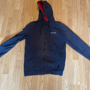 Mörkblå hoodie från Race Marine - Säljer en mörkblå hoodie från Race Marine med dragkedja och röd insida på huvan. Tröjan har långa ärmar, fickor framtill och justerbar huva med snören. Perfekt för en avslappnad stil.
