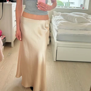 Neo Noir Satinkjol - Supersnygg långkjol i champagnefärgad satin från Neo Noir. Kjolen heter ’bovary skirt’ och säljs för 350kr pga slitningar (skriv gärna för fler bilder!). Kjolen är M men passar som lowwaist för mindre storlekar!Perfekt kjol för sommaren!! 💗